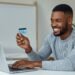 Young adult holding a new credit card after approval, smiling at laptop screen